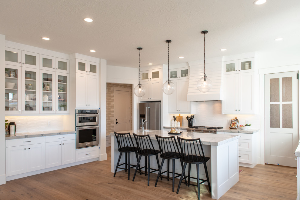 Photo of a large transitional u-shaped separate kitchen in Salt Lake City with a farmhouse sink, shaker cabinets, white cabinets, marble benchtops, white splashback, porcelain splashback, stainless steel appliances, light hardwood floors, with island, beige floor and grey benchtop.