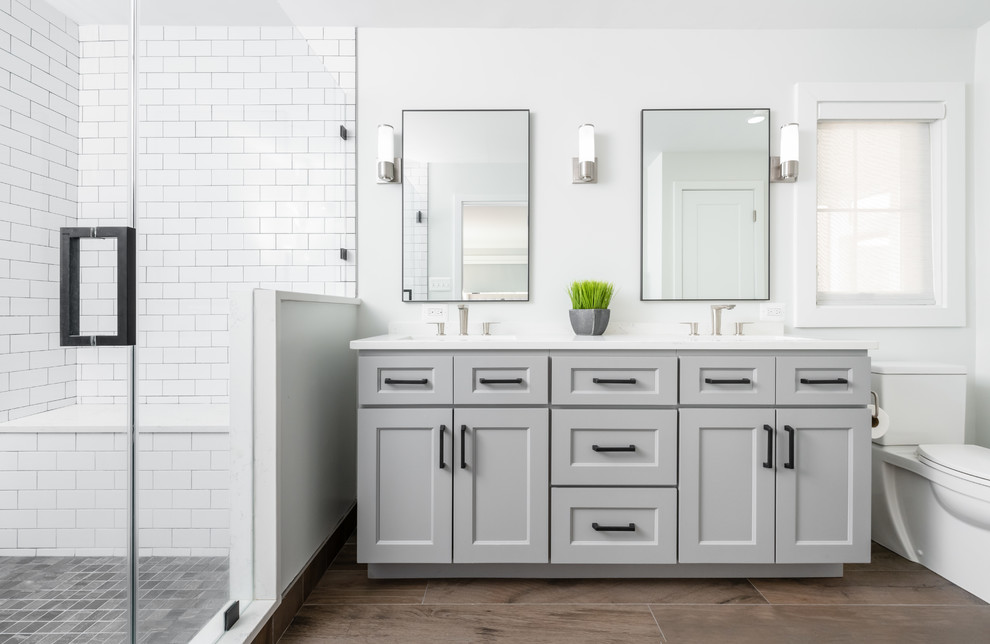 Photo of a mid-sized transitional 3/4 bathroom in DC Metro with recessed-panel cabinets, grey cabinets, an alcove shower, a two-piece toilet, white tile, subway tile, white walls, an undermount sink, brown floor, a hinged shower door, white benchtops and engineered quartz benchtops.