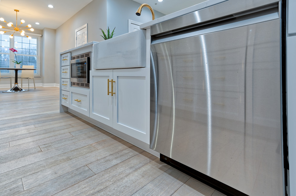 Photo of a mid-sized contemporary galley eat-in kitchen in Philadelphia with a farmhouse sink, shaker cabinets, white cabinets, quartz benchtops, white splashback, glass tile splashback, stainless steel appliances, light hardwood floors, with island, beige floor and white benchtop.