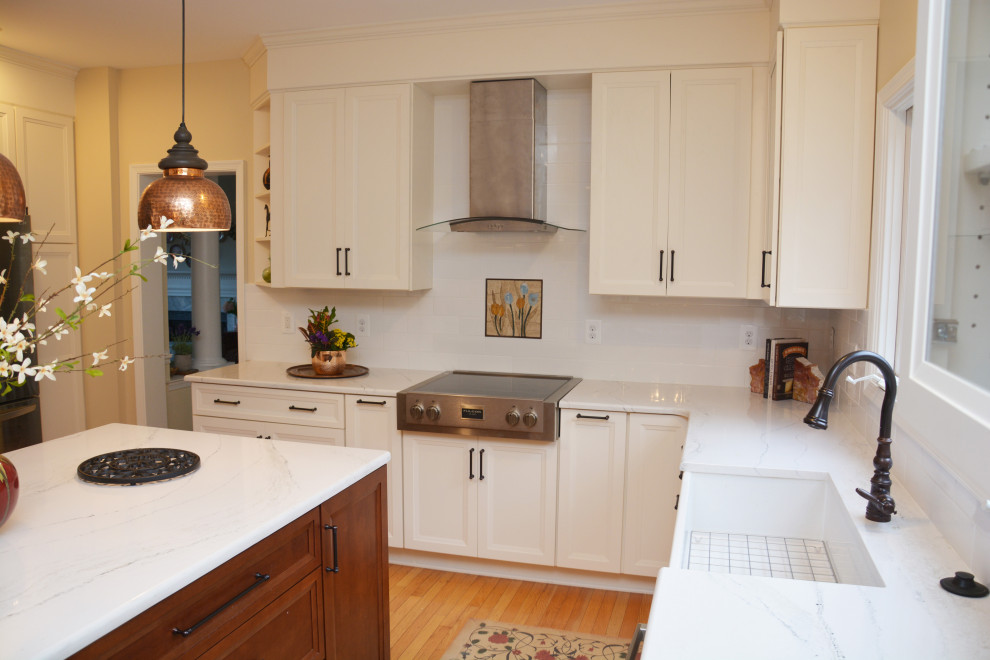 Design ideas for a mid-sized transitional l-shaped open plan kitchen in DC Metro with a farmhouse sink, recessed-panel cabinets, white cabinets, quartz benchtops, white splashback, stainless steel appliances, light hardwood floors, with island, brown floor and white benchtop.