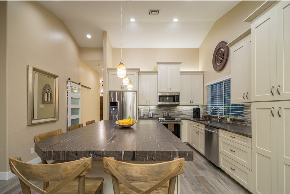 This is an example of a mid-sized transitional l-shaped open plan kitchen in Phoenix with an undermount sink, shaker cabinets, white cabinets, granite benchtops, grey splashback, stone tile splashback, stainless steel appliances, with island, brown floor, brown benchtop and laminate floors.