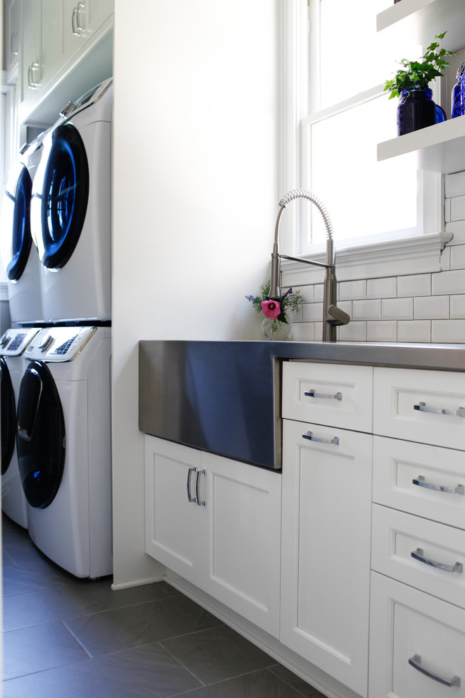 Design ideas for a transitional single-wall dedicated laundry room in Raleigh with a farmhouse sink, recessed-panel cabinets, white cabinets, stainless steel benchtops, white walls, a stacked washer and dryer, grey floor and grey benchtop.