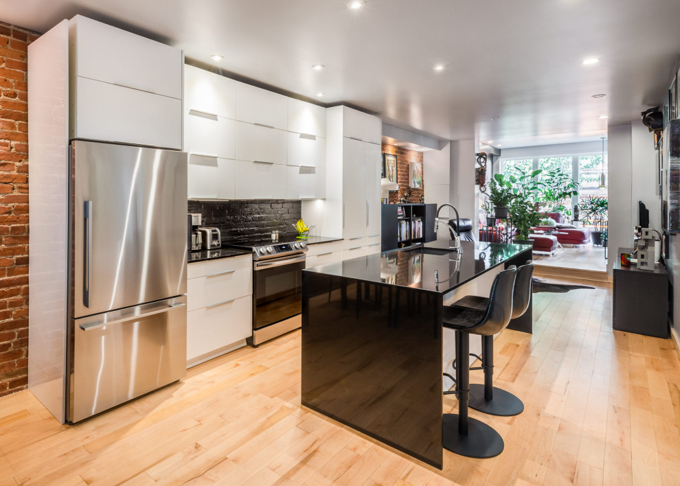 Large contemporary open plan kitchen in Montreal with white cabinets, medium hardwood flooring, an island, brown floors, black worktops, black splashback and brick splashback.