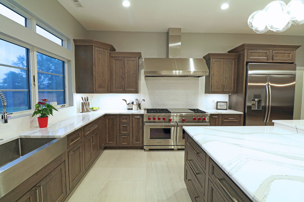 This is an example of a large transitional u-shaped eat-in kitchen in Boston with a farmhouse sink, beaded inset cabinets, brown cabinets, quartz benchtops, white splashback, porcelain splashback, stainless steel appliances, porcelain floors, with island, beige floor and white benchtop.