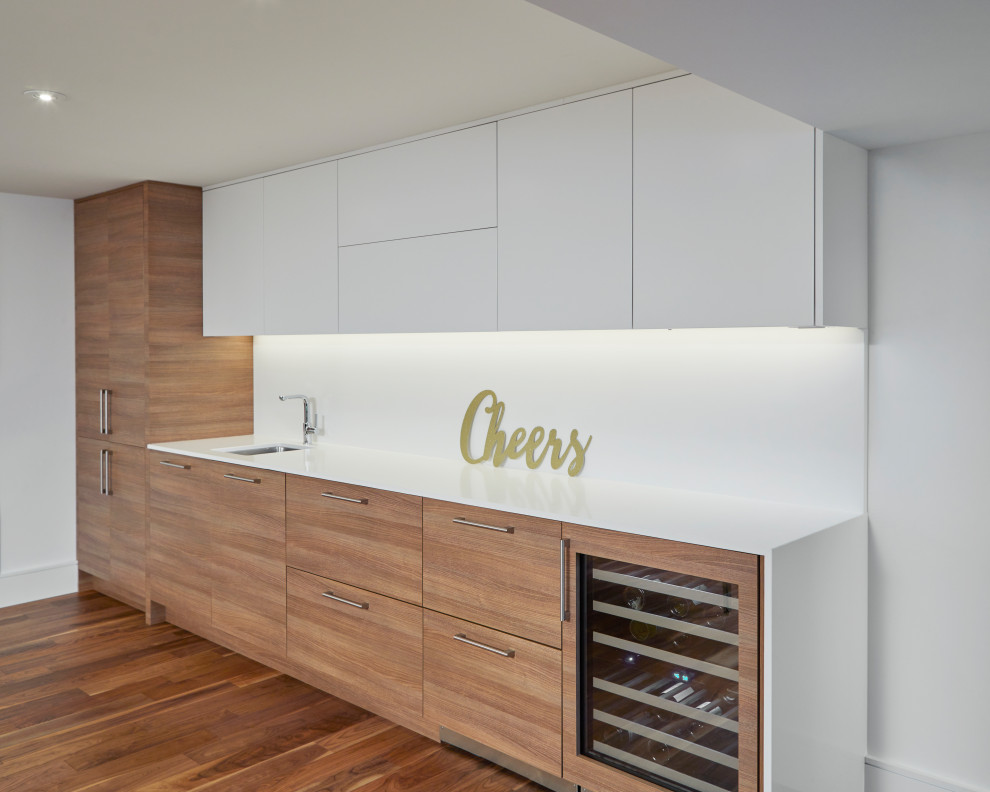 This is an example of a mid-sized contemporary galley wet bar in Toronto with an undermount sink, flat-panel cabinets, white cabinets, quartz benchtops, white splashback, stone slab splashback, medium hardwood floors, brown floor and white benchtop.