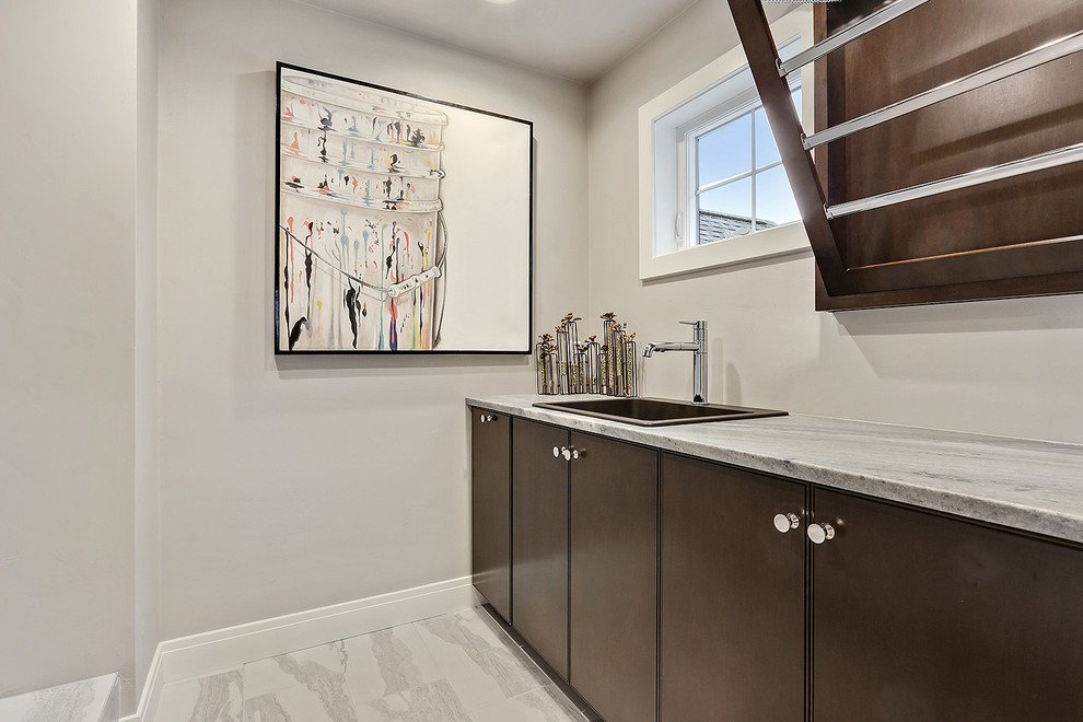 Inspiration for a mid-sized beach style dedicated laundry room in Other with a drop-in sink, flat-panel cabinets, brown cabinets, marble benchtops, grey walls, ceramic floors, a side-by-side washer and dryer, white floor and blue benchtop.