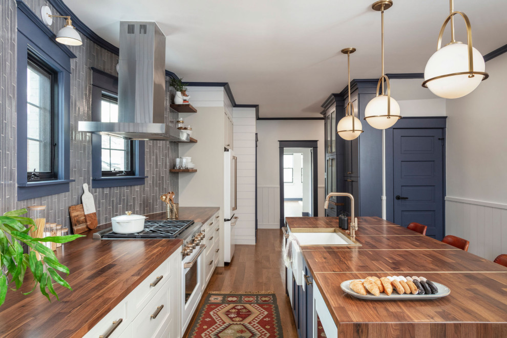 Large transitional single-wall eat-in kitchen in Denver with a farmhouse sink, shaker cabinets, white cabinets, wood benchtops, grey splashback, porcelain splashback, white appliances, medium hardwood floors, with island, brown floor, brown benchtop and recessed.