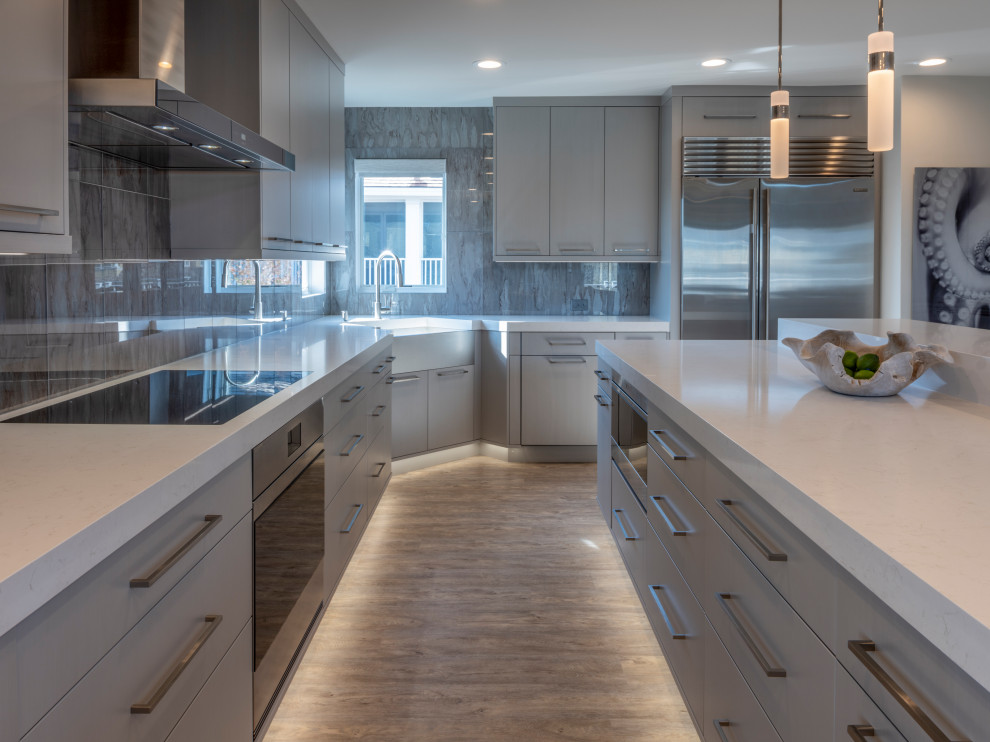 Photo of a large transitional l-shaped eat-in kitchen in New York with a drop-in sink, flat-panel cabinets, grey cabinets, quartz benchtops, blue splashback, glass tile splashback, stainless steel appliances, light hardwood floors, with island, grey floor and white benchtop.