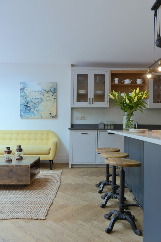 Photo of a mid-sized traditional l-shaped open plan kitchen in Gloucestershire with a farmhouse sink, flat-panel cabinets, white cabinets, solid surface benchtops, white splashback, glass sheet splashback, black appliances, light hardwood floors, with island, brown floor and grey benchtop.