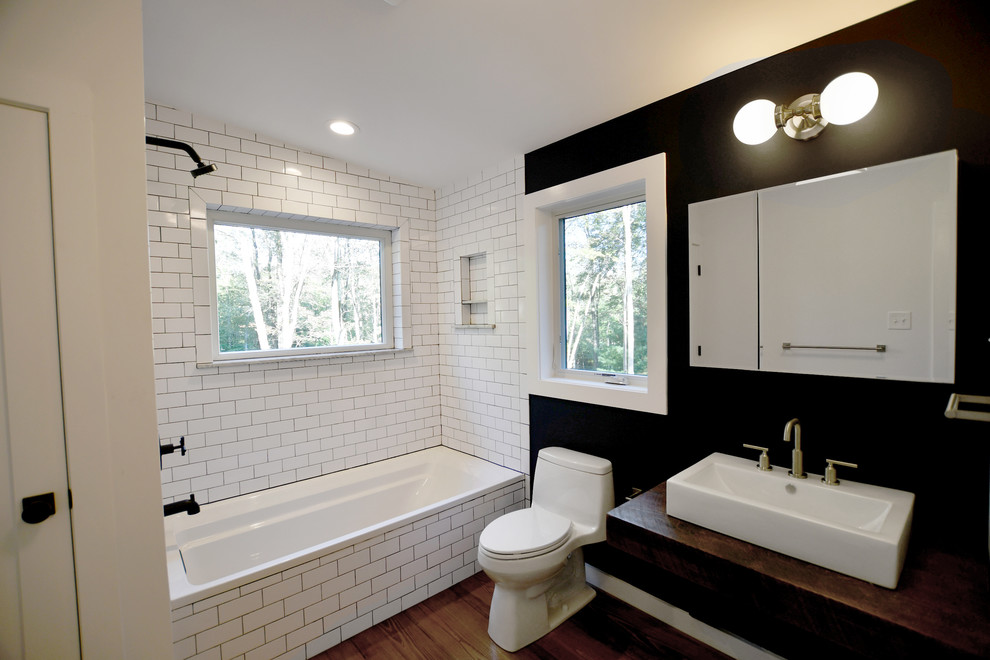 Mid-sized midcentury master bathroom in New York with open cabinets, a drop-in tub, a shower/bathtub combo, a one-piece toilet, white tile, subway tile, black walls, medium hardwood floors, wood benchtops, brown floor, an open shower and brown benchtops.