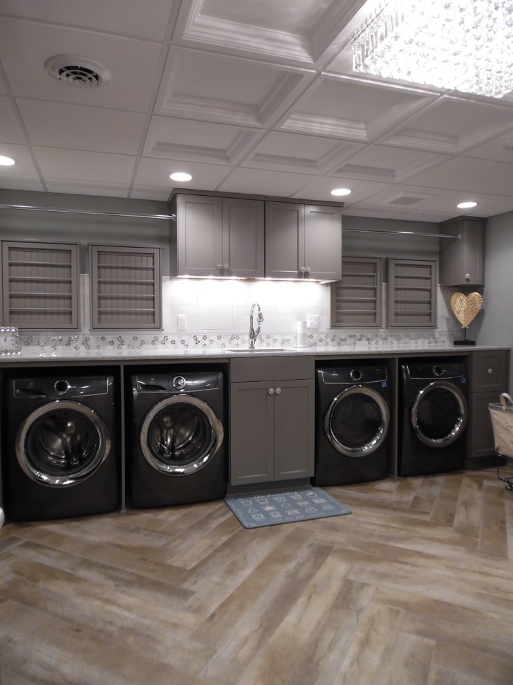 Photo of a large traditional utility room in Milwaukee with an undermount sink, recessed-panel cabinets, grey cabinets, quartz benchtops, vinyl floors, a side-by-side washer and dryer, brown floor and white benchtop.