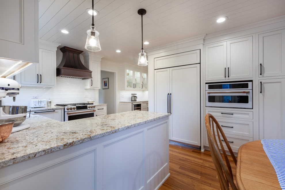 This is an example of a mid-sized country l-shaped eat-in kitchen in Chicago with a farmhouse sink, beaded inset cabinets, white cabinets, granite benchtops, white splashback, porcelain splashback, white appliances, light hardwood floors, a peninsula, orange floor, beige benchtop and timber.