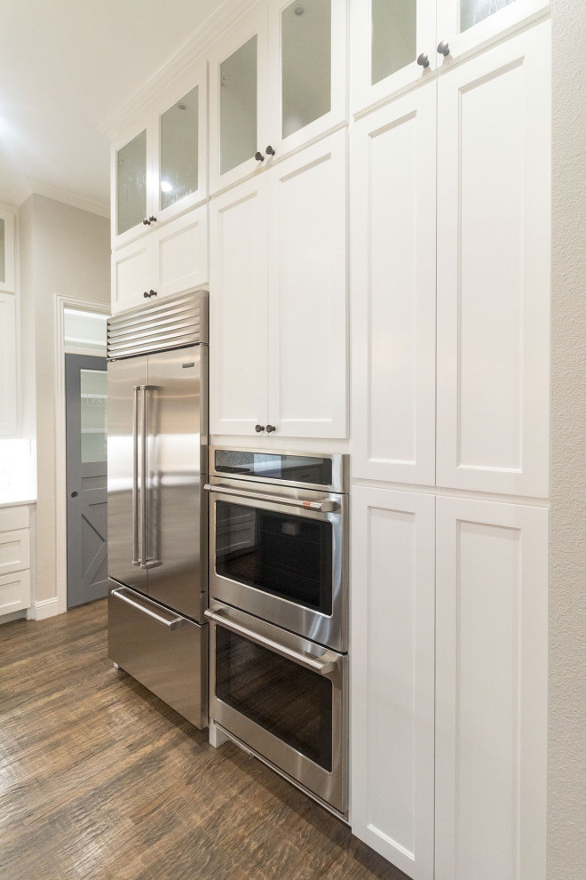 Large transitional l-shaped medium tone wood floor and brown floor open concept kitchen photo in Dallas with a farmhouse sink, shaker cabinets, white cabinets, quartz countertops, white backsplash, mosaic tile backsplash, stainless steel appliances, an island and white countertops