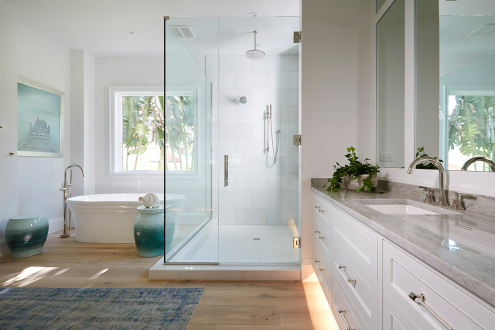 This is an example of a beach style bathroom in Miami with shaker cabinets, white cabinets, a freestanding tub, a corner shower, white tile, white walls, medium hardwood floors, an undermount sink, brown floor and a hinged shower door.