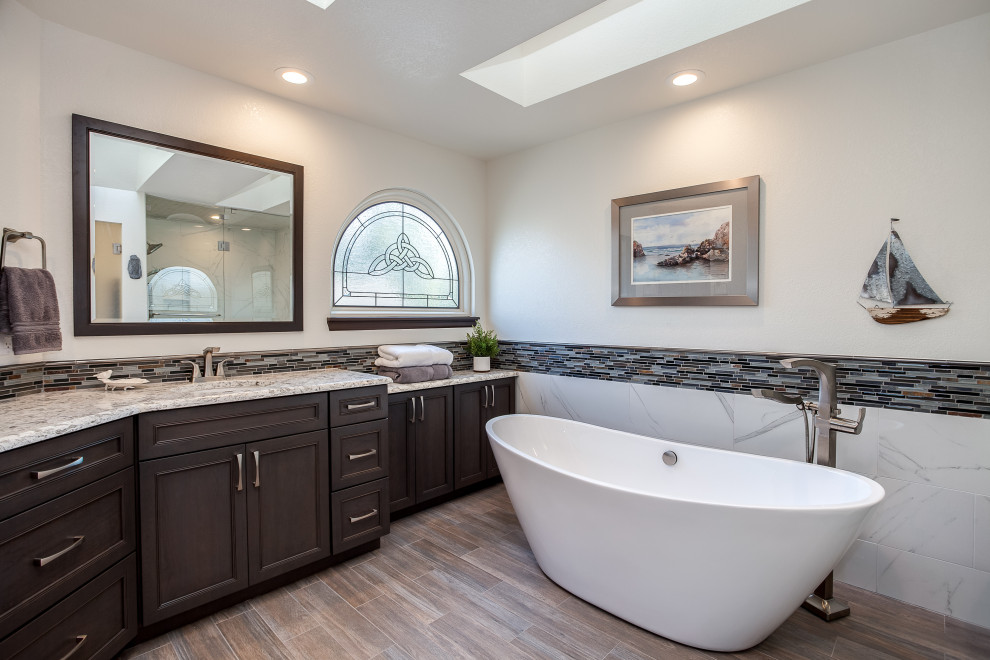Photo of a large contemporary master bathroom in Denver with shaker cabinets, brown cabinets, a japanese tub, a curbless shower, gray tile, white walls, medium hardwood floors, an integrated sink, granite benchtops, brown floor, a hinged shower door, white benchtops, a shower seat, a double vanity and a built-in vanity.