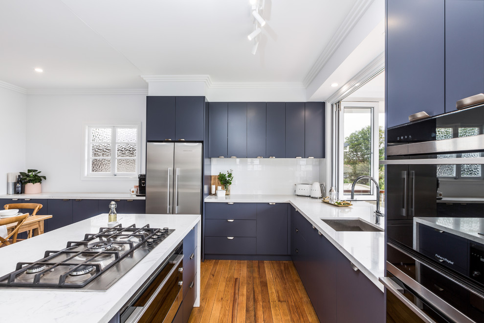 Inspiration for a small transitional l-shaped eat-in kitchen in Brisbane with an undermount sink, flat-panel cabinets, blue cabinets, quartz benchtops, white splashback, subway tile splashback, black appliances, medium hardwood floors, with island, brown floor and white benchtop.