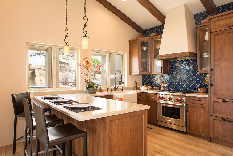 Inspiration for a mediterranean u-shaped kitchen in Los Angeles with a farmhouse sink, glass-front cabinets, medium wood cabinets, blue splashback, stainless steel appliances, light hardwood floors, a peninsula and beige floor.