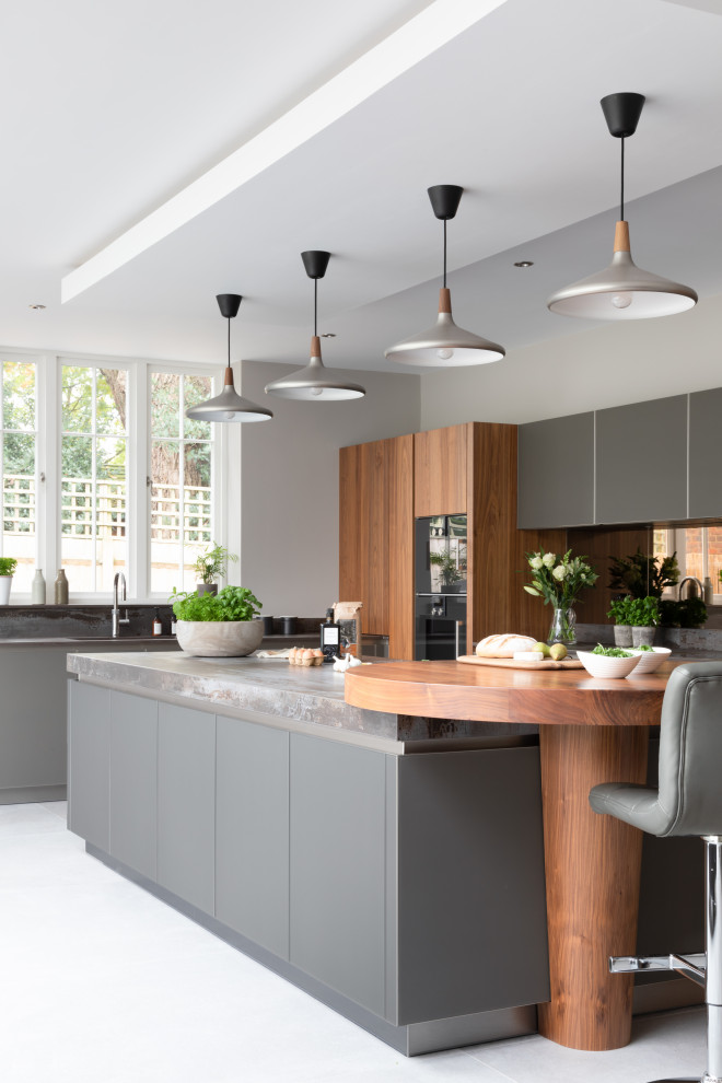 This is an example of a large contemporary open plan kitchen in Hampshire with an integrated sink, glass-front cabinets, grey cabinets, quartzite benchtops, metallic splashback, mirror splashback, black appliances, with island and grey benchtop.