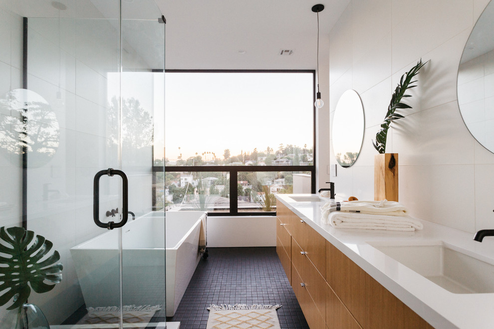 This is an example of a mid-sized contemporary master bathroom in Los Angeles with flat-panel cabinets, light wood cabinets, a freestanding tub, a corner shower, a one-piece toilet, white tile, porcelain tile, white walls, mosaic tile floors, an undermount sink, quartzite benchtops, black floor, a hinged shower door and white benchtops.