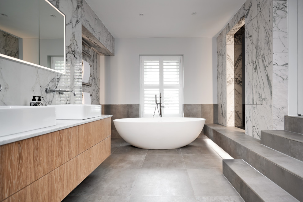 Photo of a large contemporary ensuite bathroom in Other with flat-panel cabinets, light wood cabinets, a freestanding bath, grey tiles, marble tiles, ceramic flooring, grey floors, a feature wall, double sinks, a floating vanity unit, white walls, a vessel sink and white worktops.