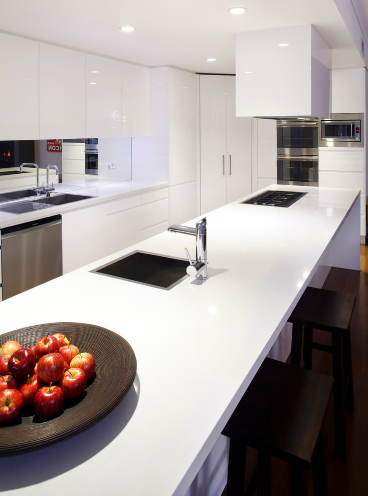 This is an example of a mid-sized modern l-shaped eat-in kitchen in Sydney with a drop-in sink, flat-panel cabinets, white cabinets, glass benchtops, mirror splashback, medium hardwood floors, with island, metallic splashback and stainless steel appliances.