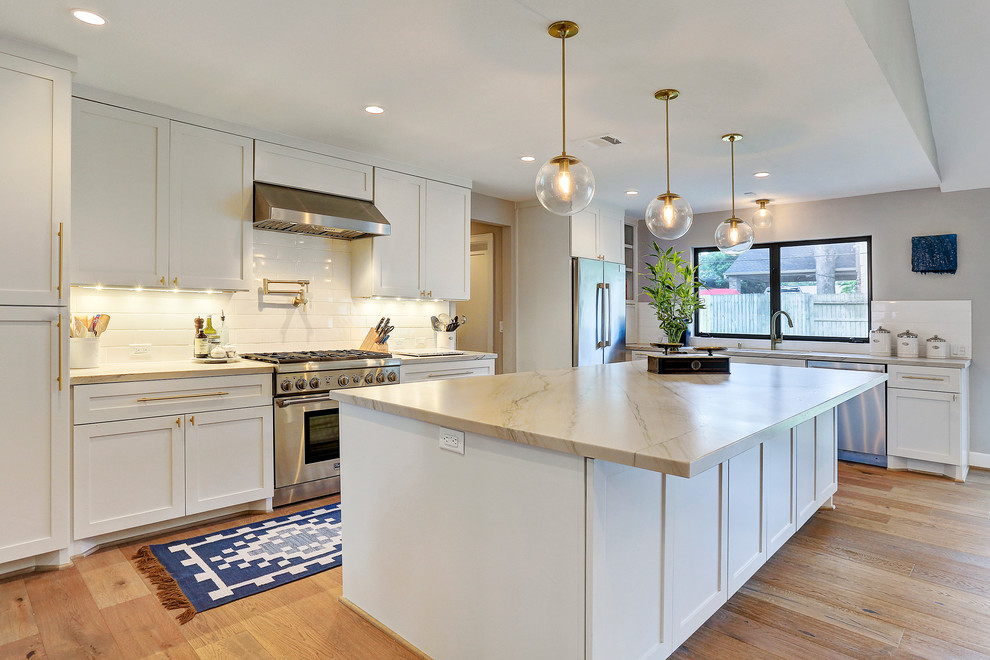 Mid-sized transitional l-shaped eat-in kitchen in Houston with a double-bowl sink, shaker cabinets, white cabinets, marble benchtops, white splashback, ceramic splashback, stainless steel appliances, light hardwood floors and with island.