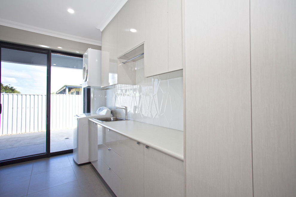 Photo of a mid-sized modern single-wall dedicated laundry room in Other with a single-bowl sink, laminate benchtops, white walls, flat-panel cabinets, white cabinets, white splashback, glass sheet splashback, ceramic floors, a stacked washer and dryer, grey floor and white benchtop.