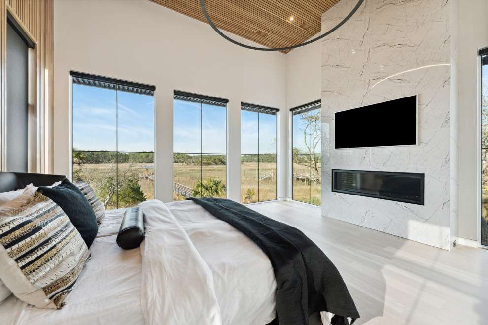 Modelo de dormitorio principal moderno grande con paredes blancas, suelo de madera clara, chimenea lineal, marco de chimenea de piedra, madera y panelado