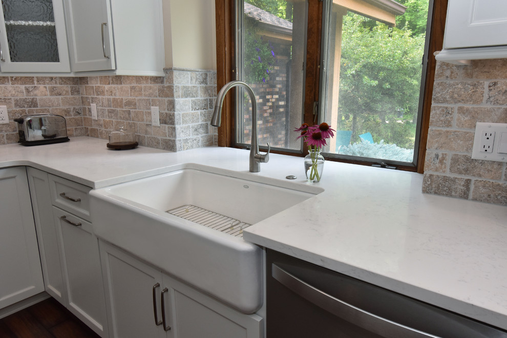 Mid-sized transitional galley separate kitchen in Milwaukee with a farmhouse sink, recessed-panel cabinets, white cabinets, quartz benchtops, beige splashback, travertine splashback, stainless steel appliances, vinyl floors, with island, brown floor and white benchtop.