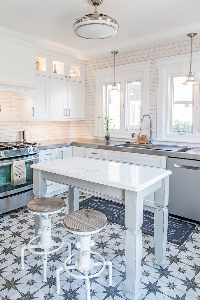 Country l-shaped kitchen in Philadelphia with an integrated sink, shaker cabinets, white cabinets, stainless steel benchtops, white splashback, subway tile splashback, stainless steel appliances, with island and multi-coloured floor.