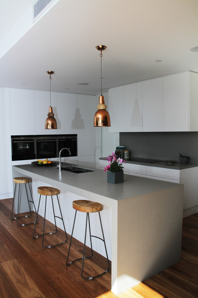 Contemporary kitchen in Sydney with a drop-in sink, white cabinets, solid surface benchtops, grey splashback, stone slab splashback, black appliances, dark hardwood floors, with island and grey benchtop.