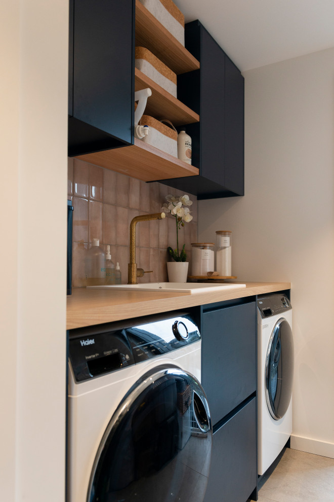 Design ideas for a mid-sized beach style single-wall dedicated laundry room in Auckland with a drop-in sink, flat-panel cabinets, blue cabinets, laminate benchtops, pink splashback, porcelain splashback, white walls, porcelain floors, a side-by-side washer and dryer, grey floor and brown benchtop.