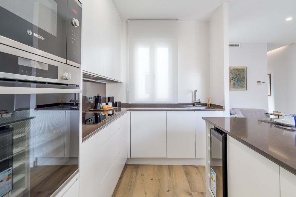 Inredning av ett modernt litet grå grått kök och matrum, med en enkel diskho, släta luckor, vita skåp, bänkskiva i kalksten, grått stänkskydd, stänkskydd i kalk, rostfria vitvaror, mellanmörkt trägolv, en köksö och brunt golv