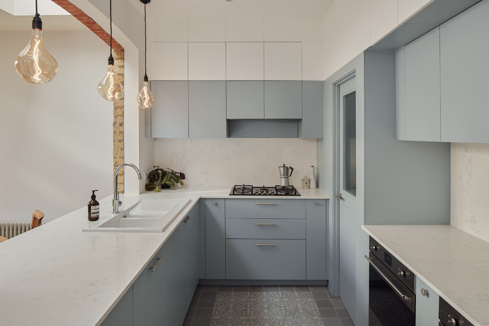 Photo of a scandinavian kitchen in London with a drop-in sink, flat-panel cabinets, blue cabinets, white splashback, stone slab splashback, stainless steel appliances, a peninsula, grey floor and white benchtop.