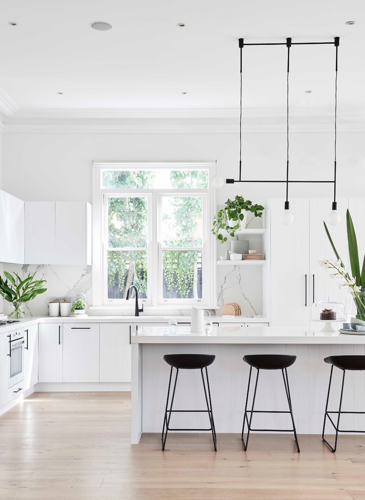 This is an example of a large modern l-shaped eat-in kitchen in Melbourne with an undermount sink, white cabinets, quartz benchtops, white splashback, porcelain splashback, white appliances, light hardwood floors, with island, brown floor and white benchtop.
