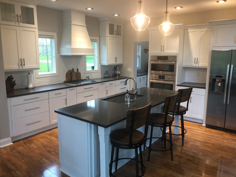 Laminate floor and brown floor kitchen photo with a single-bowl sink, shaker cabinets, white cabinets, quartzite countertops, white backsplash, subway tile backsplash, stainless steel appliances, an island and gray countertops