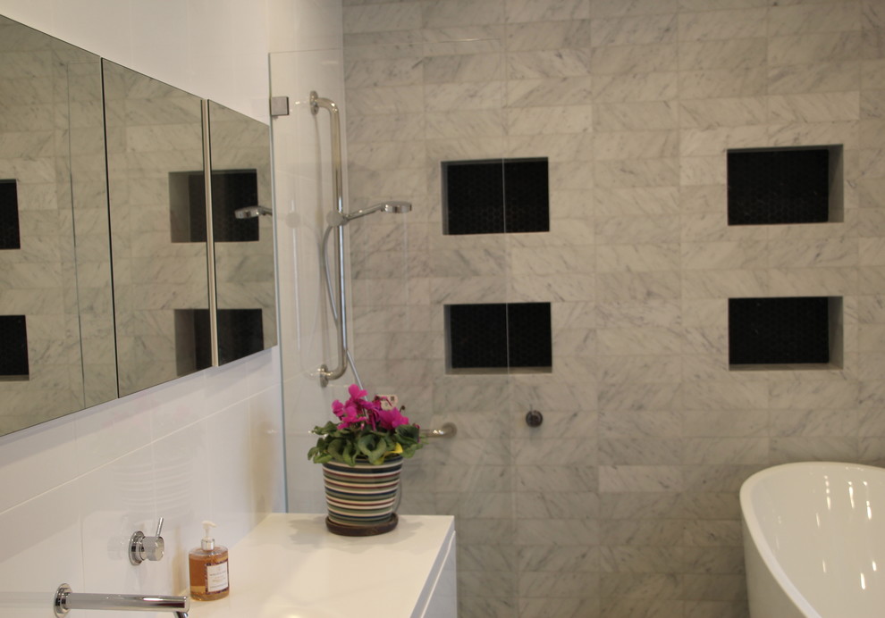 This is an example of a mid-sized contemporary kids bathroom in Sydney with glass-front cabinets, white cabinets, a freestanding tub, an open shower, a wall-mount toilet, black and white tile, marble, white walls, porcelain floors, an integrated sink, solid surface benchtops, grey floor and an open shower.