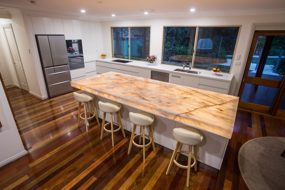Photo of a large contemporary kitchen pantry in Brisbane with a double-bowl sink, flat-panel cabinets, white cabinets, quartzite benchtops, black appliances, dark hardwood floors, with island, brown floor and brown benchtop.