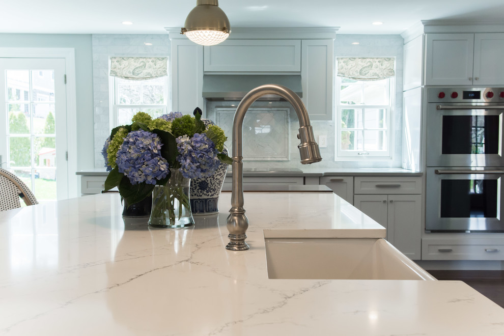Large transitional l-shaped eat-in kitchen in Providence with a farmhouse sink, shaker cabinets, white cabinets, quartz benchtops, white splashback, marble splashback, stainless steel appliances, medium hardwood floors, with island, brown floor and white benchtop.