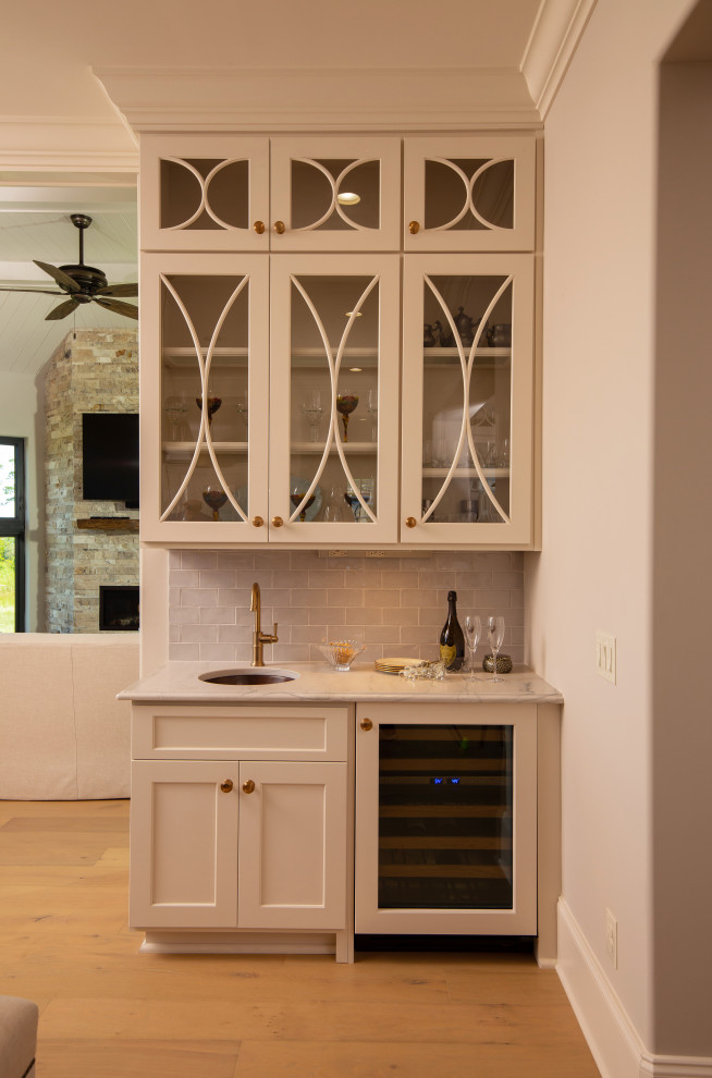 Photo of a small traditional single-wall wet bar in Other with an undermount sink, glass-front cabinets, beige cabinets, quartzite benchtops, grey splashback, glass tile splashback, light hardwood floors, beige floor and grey benchtop.