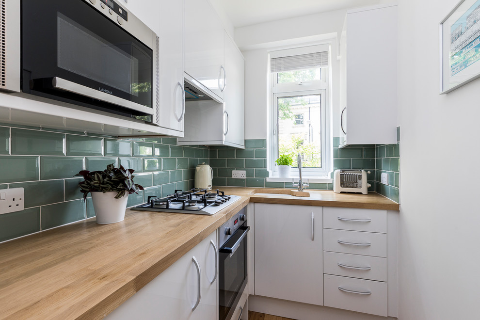 Small contemporary l-shaped kitchen in London with an undermount sink, flat-panel cabinets, white cabinets, wood benchtops, blue splashback, subway tile splashback, stainless steel appliances and no island.