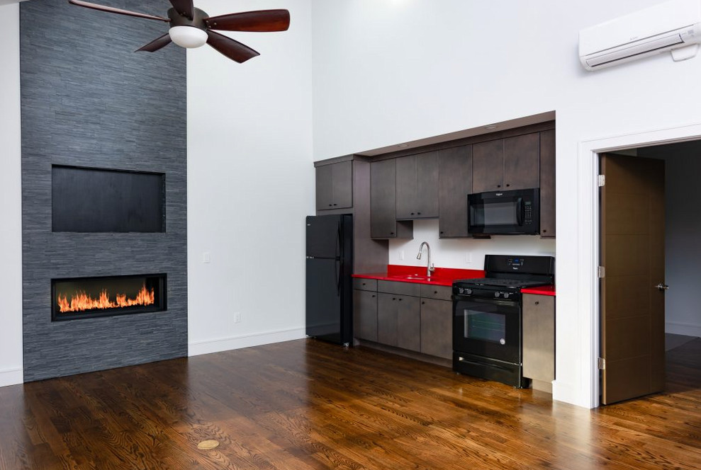 Small transitional single-wall medium tone wood floor and brown floor open concept kitchen photo with an undermount sink, flat-panel cabinets, dark wood cabinets, solid surface countertops, white backsplash, black appliances, no island and red countertops