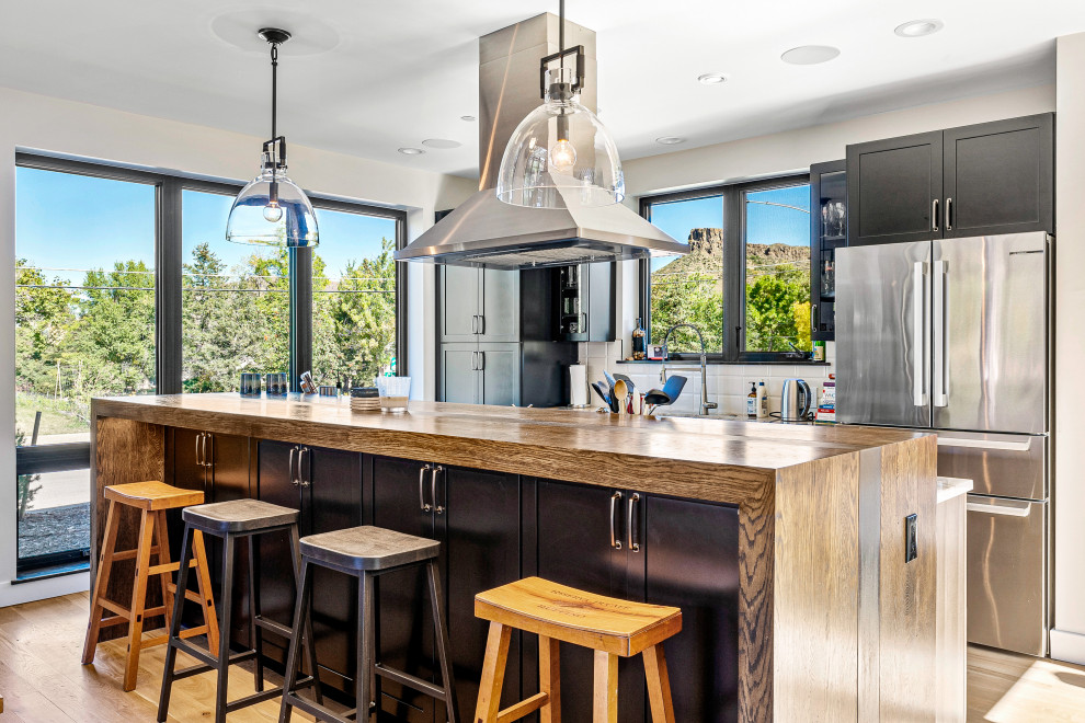 Example of a transitional galley light wood floor and beige floor kitchen design in Denver with shaker cabinets, black cabinets, wood countertops, white backsplash, stainless steel appliances, an island and brown countertops