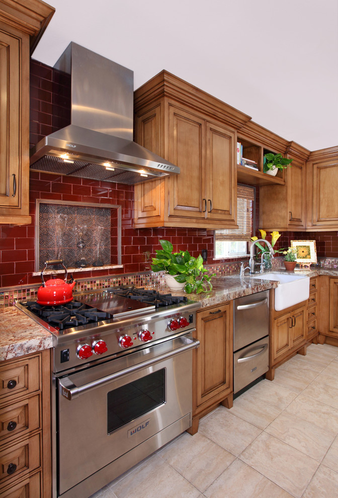 Photo of a country kitchen in Los Angeles with a farmhouse sink, granite benchtops, red splashback, glass tile splashback, stainless steel appliances, raised-panel cabinets, medium wood cabinets and porcelain floors.