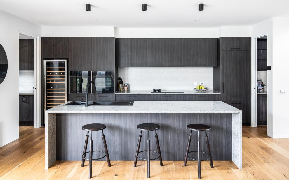 Photo of a large contemporary galley kitchen in Melbourne with a double-bowl sink, flat-panel cabinets, dark wood cabinets, white splashback, subway tile splashback, panelled appliances, light hardwood floors, with island, beige floor and grey benchtop.