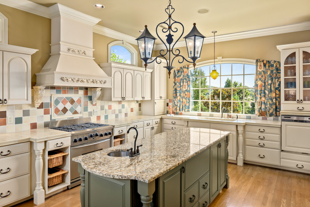 This is an example of a traditional l-shaped kitchen in Nashville with an undermount sink, raised-panel cabinets, white cabinets, tile benchtops, multi-coloured splashback, stainless steel appliances, medium hardwood floors, with island, brown floor and beige benchtop.