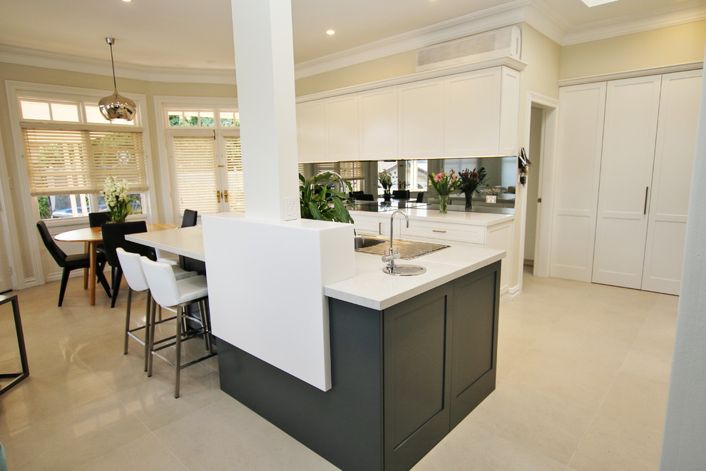 This is an example of a large transitional l-shaped eat-in kitchen in Sydney with a drop-in sink, shaker cabinets, white cabinets, quartz benchtops, grey splashback, glass sheet splashback, stainless steel appliances, ceramic floors and with island.