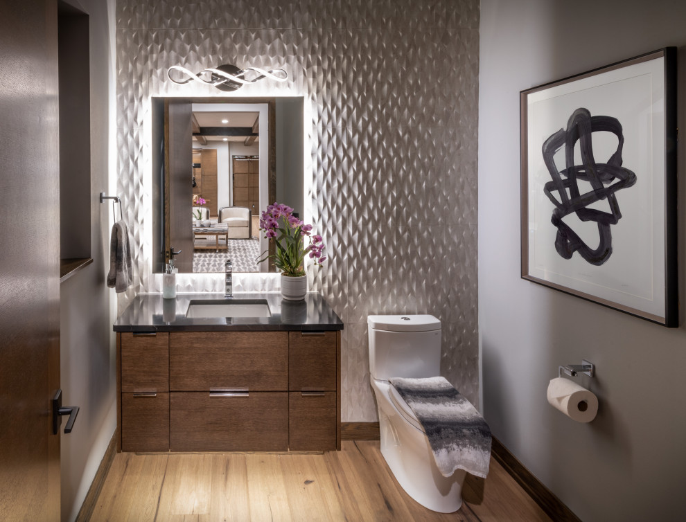 Photo of a small modern cloakroom in Milwaukee with flat-panel cabinets, brown cabinets, a two-piece toilet, grey tiles, ceramic tiles, grey walls, medium hardwood flooring, a submerged sink, granite worktops, brown floors, black worktops and a floating vanity unit.
