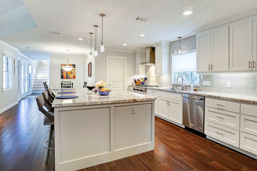 Photo of a mid-sized traditional single-wall open plan kitchen in Houston with an undermount sink, shaker cabinets, white cabinets, granite benchtops, grey splashback, subway tile splashback, stainless steel appliances, medium hardwood floors, with island and brown floor.