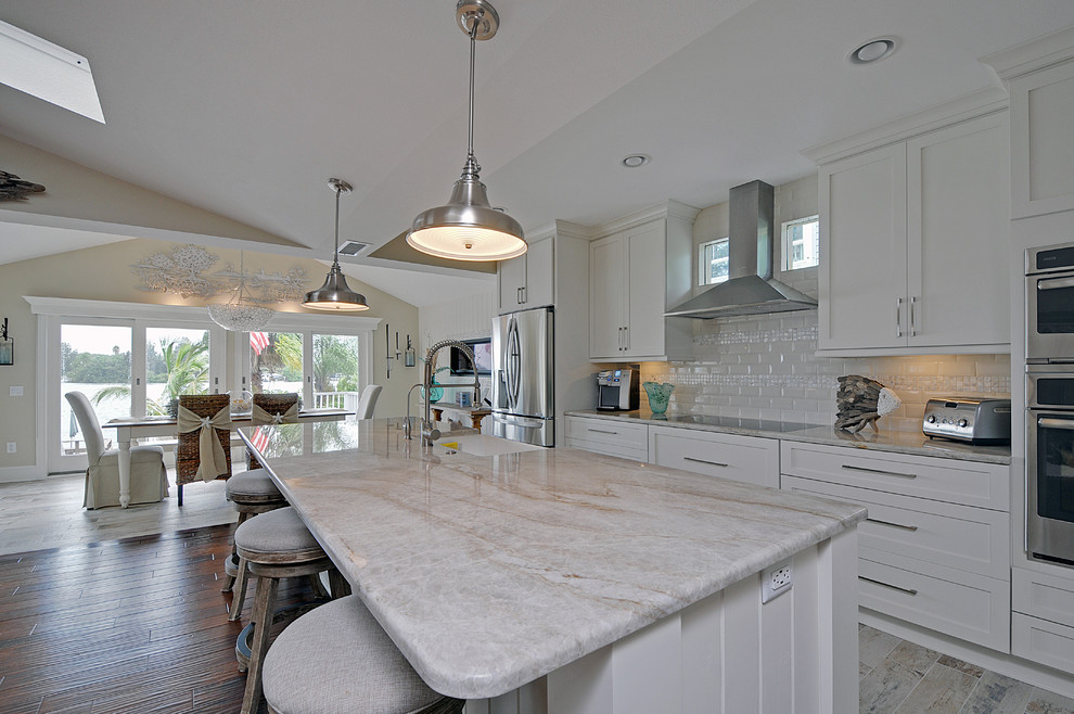 Mid-sized beach style single-wall open plan kitchen in Tampa with a farmhouse sink, shaker cabinets, white cabinets, granite benchtops, white splashback, ceramic splashback, stainless steel appliances, vinyl floors and with island.